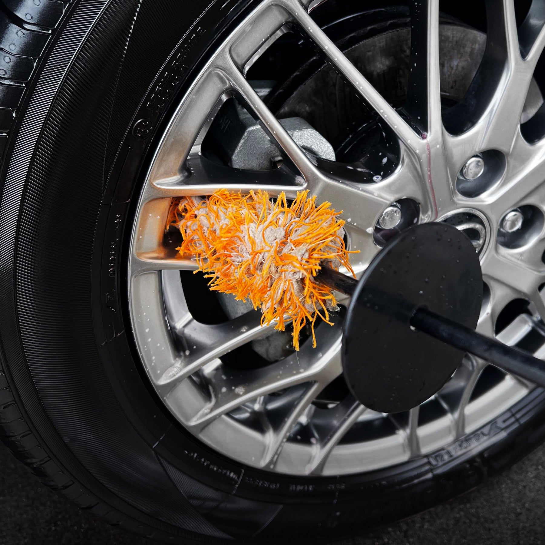 pazzola CARPRO Flat & Spot Brush con setole arancioni mentre pulisce un cerchione di un'auto. Le setole morbide sono in azione tra i raggi del cerchione, rimuovendo sporco e residui.
