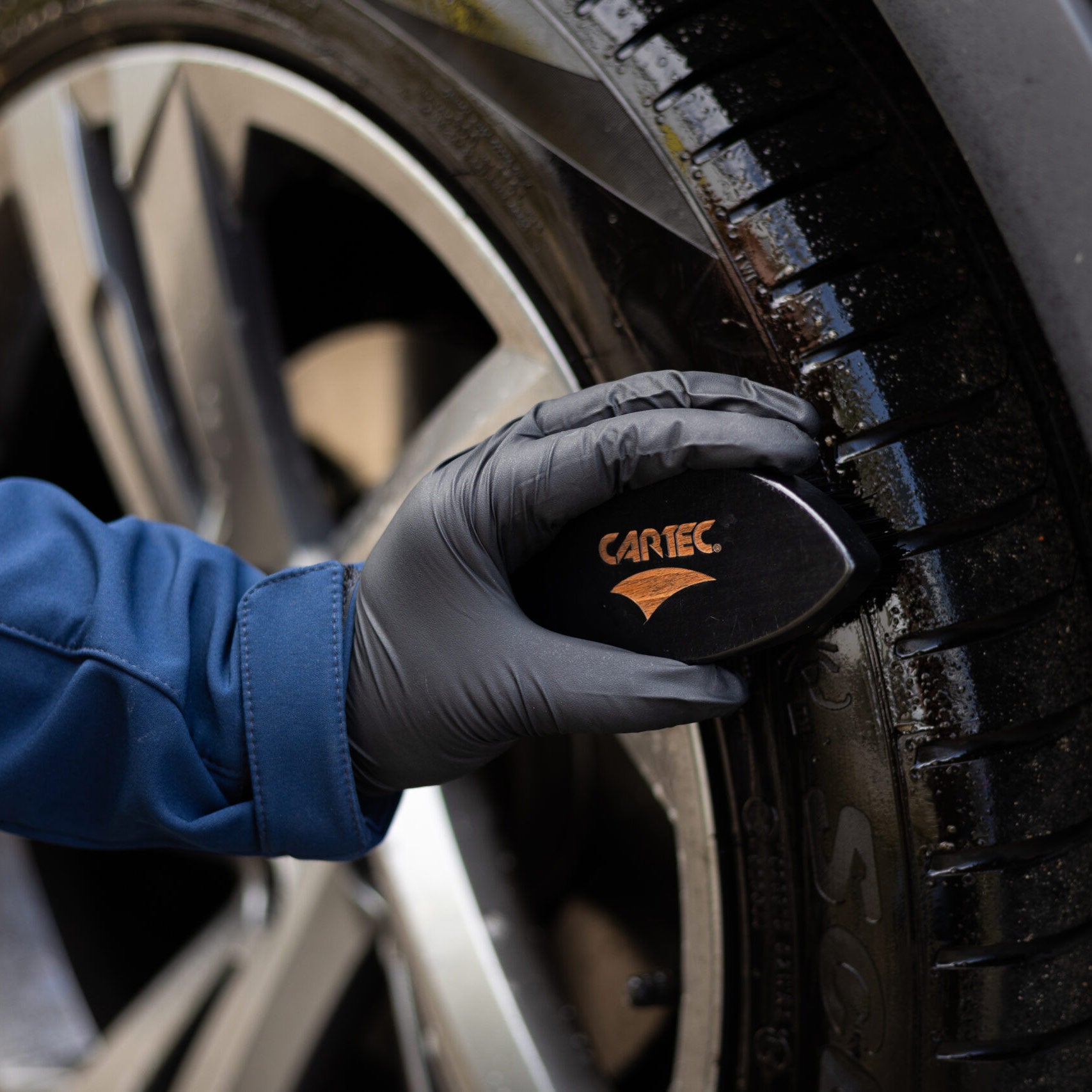 Mano con guanto nero che utilizza la spazzola CARTEC Dynamic Tyre Brush per pulire un pneumatico.